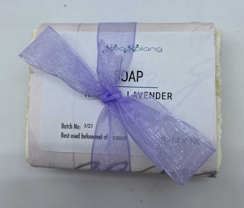 A natural coloured soap in an envelope of lavender and white paper with a label and a lavender ribbon tied in a bow