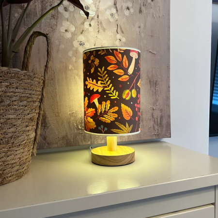 Autumn-themed table lamp with a wooden base and a warm forest pattern on the lampshade, placed on a white unit beside a woven basket and wall art.