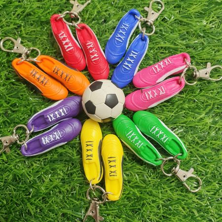 Football boots keyrings in blue, pink, green, yellow, purple, orange and red, surrounding a white & black soccer ball, all laid on the grass.