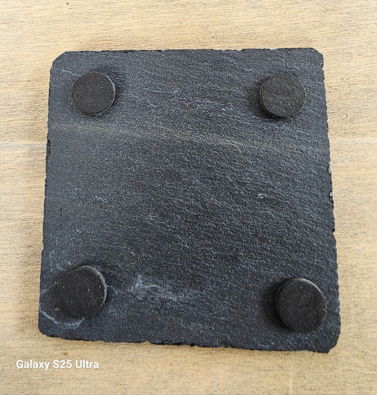 Square slate coaster with four black rubber feet on a wooden surface