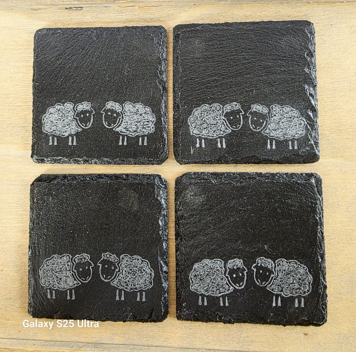 Four slate coasters with sheep illustrations on a wooden board.