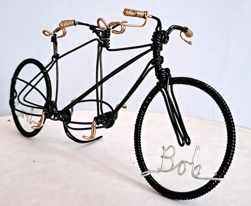 Black miniature tandem sculpture made from galvanised steel and hand painted black with gold handles, seat and pedals. Made as a wedding cake topper and personalised with two names written in wire and set into the front and back wheel of the bicycle