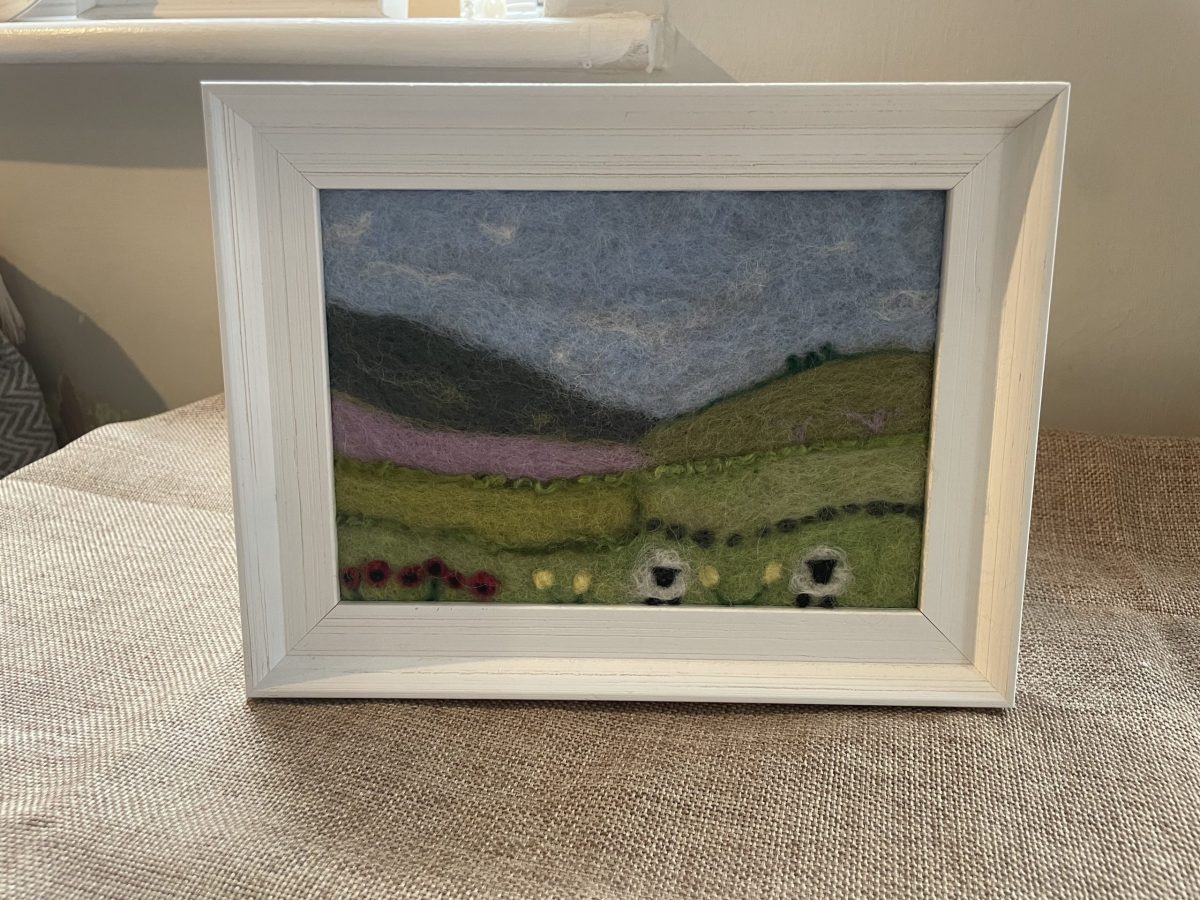 A needle-felted wool landscape of green hills and sheep in a white frame